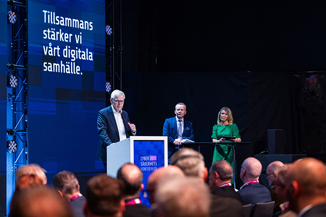 Man i talarstol, man och kvinna i bakgrunden